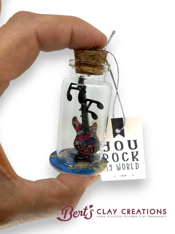 Glass bottle with cork stopper containing a miniature guitar inside, held by a hand. Tag with 'You Rock My World' text and 'Bert's Clay Creations' branding.